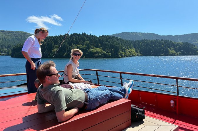 Travelers on the open deck of the Lake Ashi boat, looking across the water toward a forested shore